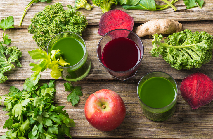 deux verres de jus de fruits vert et un verre de jus de fruits rouge foncé (betterave) sur un fond en bois, avec des morceaux de salade, une coupe transversale de betterave, des branches de céleri, du gingembre, du persil, une pomme et du chou frisé en guise de garniture. Vue en plongée.