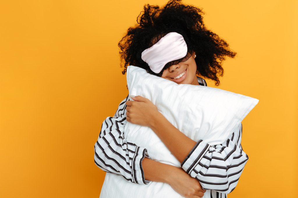 Dormir. Rêves. Portrait de femme. Une fille afro-américaine en pyjama et masque de sommeil serre un oreiller dans ses bras et sourit, sur un fond jaune.